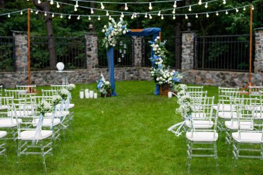 Açık havada düğün için bir yer. Misafirler için oturma alanı var. Sandalyelerle süslenmiş. Mavi kumaş, çiçek aranjmanı ve mumlarla süslenmiş ahşap bir kemer. Yumuşak seçici odak.