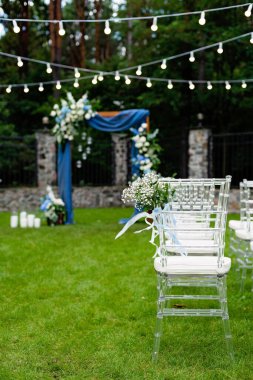 Açık havada düğün için bir yer. Misafirler için oturma alanı var. Sandalyelerle süslenmiş. Mavi kumaş, çiçek aranjmanı ve mumlarla süslenmiş ahşap bir kemer. Yumuşak seçici odak.