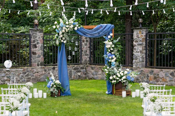 Açık havada düğün için bir yer. Misafirler için oturma alanı var. Sandalyelerle süslenmiş. Mavi kumaş, çiçek aranjmanı ve mumlarla süslenmiş ahşap bir kemer. Yumuşak seçici odak.