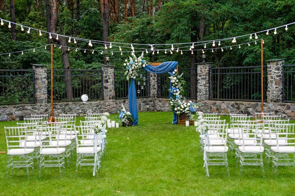 Açık havada düğün için bir yer. Misafirler için oturma alanı var. Sandalyelerle süslenmiş. Mavi kumaş, çiçek aranjmanı ve mumlarla süslenmiş ahşap bir kemer. Yumuşak seçici odak.