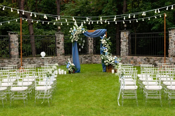 Açık havada düğün için bir yer. Misafirler için oturma alanı var. Sandalyelerle süslenmiş. Mavi kumaş, çiçek aranjmanı ve mumlarla süslenmiş ahşap bir kemer. Yumuşak seçici odak.
