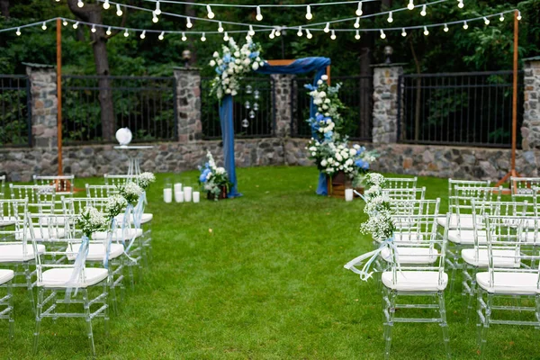 Açık havada düğün için bir yer. Misafirler için oturma alanı var. Sandalyelerle süslenmiş. Mavi kumaş, çiçek aranjmanı ve mumlarla süslenmiş ahşap bir kemer. Yumuşak seçici odak.