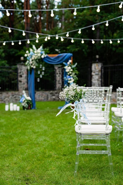 Açık havada düğün için bir yer. Misafirler için oturma alanı var. Sandalyelerle süslenmiş. Mavi kumaş, çiçek aranjmanı ve mumlarla süslenmiş ahşap bir kemer. Yumuşak seçici odak.