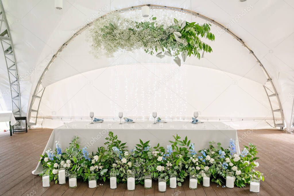 Wedding presidium in restaurant. Banquet table for newlyweds with flowers, greenery, candles and garland ligths. Lush floral arrangement. Luxury wedding decorations. Soft selective focus.