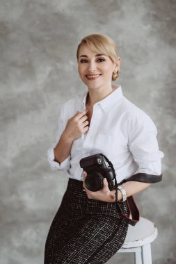 Portrait of a cheerful positive optimistic young woman photographer in the studio with a camera, on a gray background. Soft selective focus.