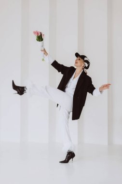 A happy stylish woman in a fashionable outfit, a light white suit with feathers, a black jacket, a baseball cap and with a bouquet of flowers in her hands is having fun. Soft selective focus.