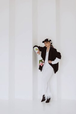 A young stylish woman in a fashionable outfit, a light white suit with feathers, a black jacket, a baseball cap and a bouquet of flowers in her hands poses on a white background. Soft selective focus.