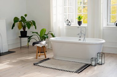 Modern banyo içerisi çok şık. Boş bir küvetin yatay görüntüsü ahşap bir zeminde, parlak bir odada, büyük bir pencerenin ve ev bitkilerinin arka planında. Yumuşak seçici odak.