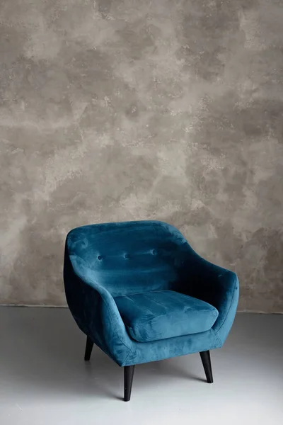 Classic blue velvet art deco armchair with wooden legs against a gray wall. Front view. Soft selective focus.