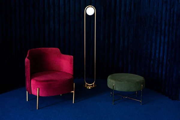 An apartment with a comfortable red velor armchair, a minimalist floor lamp and a green pouffe in a room with dark blue velvet wall panels and floor carpeting. Luxury interior design concept.