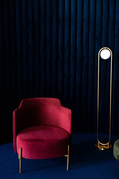 A room with a comfortable red velor armchair, a minimalist floor lamp in the living room with dark blue velvet wall panels and floor carpeting.