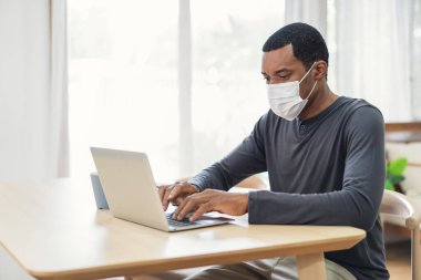 Siyah iş adamı evdeki bir masada dizüstü bilgisayarla otururken tıbbi koruyucu maske takıyor. Kendini izole etme karantinası, yeni normal. Coronavirus Covid-19 Salgını sırasında Sosyal Mesafe