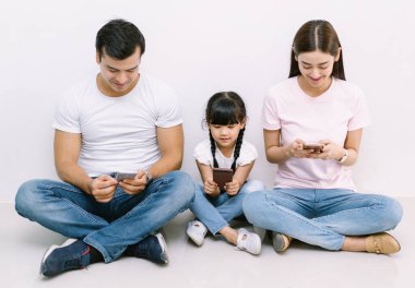 Happy technology addicted Asian family parents and kid holding smart phones having electronic devices sit on floor white background. Gadgets dependence overuse, Social media addiction concept