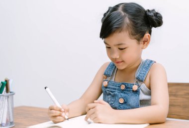 Portrait of cute little Asian girl writing or drawing, coloring pages, in notebook at desk on white background. School kid girl to do homework. Elementary school and homeschooling concept.