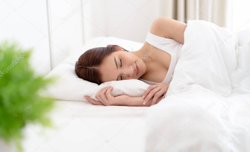 Peaceful serene beautiful young Asian woman wear pajamas lying relaxing sleeping in cozy white bed and keeping eyes closed while covered with blanket in the morning. Enjoy good healthy sleep concept.