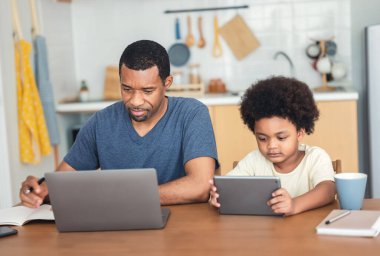Afro-Amerikan ailesi teknolojilerini kullanarak mutfaktaki masada oturuyorlar. Küçük çocuk dijital tablet kullanmayı internetten öğreniyor, video izliyor ve babam dizüstü bilgisayarla çalışıyor. Evden çalış.