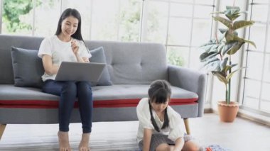 Sakin Asyalı anne dizüstü bilgisayarda çalışırken kanepeye oturur, küçük kız evde oturma odasında yerde otururken renkli plastik bloklar oynar. Evde çalışan bekar bir anne. Yeni normal kavram.