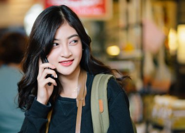 Güzel Asyalı blog yazarı sırt çantalı kadın şehirde cep telefonuyla konuşuyor. Gezgin Koreli ya da Japon kız sokak pazarında ışık saçan arka planda akıllı telefon konuşmaları yapıyor..