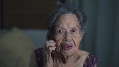 Mutlu Asyalı yaşlı kadın yatakta dinleniyor, çocuklarıyla oturma odasında akıllı telefondan konuşuyor. Teknoloji, yaşlılık ve insan konsepti