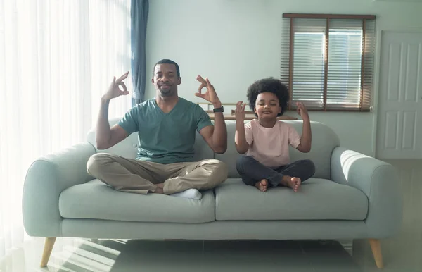 African American dad and little son calm down do practicing yoga exercises with closed eyes sit on sofa in living room. Black Family sit on couch in lotus pose position meditation together at home.