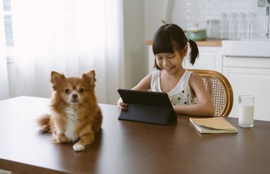 Mutlu şirin Asyalı ilkokul öğrencisi mutfakta masa başında oturmuş dijital tablet kullanarak internetten öğreniyor. Uzaktan eğitim, çocuklar tarafından kendi kendine çalışma. Eğitim ve teknoloji kavramı.