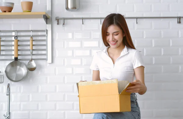 Beautiful Asian woman customer receive post shipment parcel at home. Japanese girl opening cardboard box while seated on countertop in white modern kitchen. Online shopping order, delivery concept