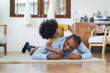 Black family have fun playing together on floor at home. Laughing African American boy carrying on Dad's back and tickling in living room at home. Black father play together with son after work.