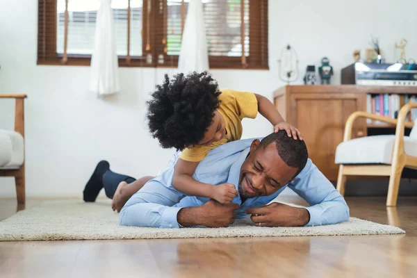 Black family have fun playing together on floor at home. Laughing African American boy carrying on Dad\'s back and tickling in living room at home. Black father play together with son after work.