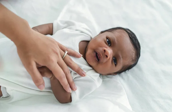 Closeup portrait of cute African american newborn baby lying on bed and looking at camera in bedroom. Mother hand lulling little child to sleeping. Child Care, Black family, Newborn baby boy. New life