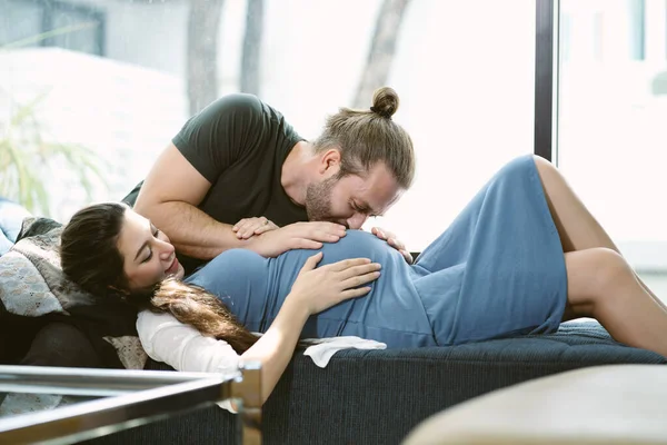 妻がソファベッドに寝そべっている間 幸せなハンサムな夫は妊娠中の妻の赤ん坊に腹でキスする 生まれていない子供への父の愛を象徴する非常に柔らかい瞬間 産前ケアの概念 — ストック写真