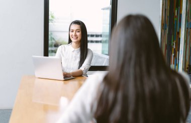 Ofiste birlikte otururken iş arkadaşlarıyla bir şeyler tartışan mutlu Asyalı kadın. Uygulamaları kullanarak dizüstü bilgisayarda çalışan dost canlısı meslektaşlar.