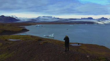 Tepede kameralı bir adamın arkasından hava panoramik görüntüsü ve buzullar ve sıradağlarla birlikte göl manzarası.