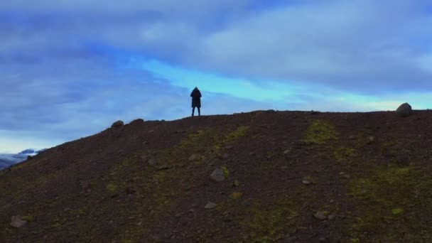 Повітряний політ позаду людини в чорній куртці на пагорбі біля озера з льодовиками та гірським масивом — стокове відео