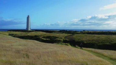Girl walking the grass path to the lighthouse in the field on a cliff of Iceland