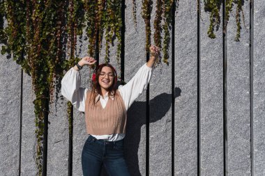 Çekici, çekici, hayalperest, neşeli, neşeli, kızıl saçlı kadın arka planda bitkilerle sokakta dans ederken kırmızı kulaklıklı müzik dinliyor. Metin için alanı kopyala