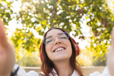 Gözlüklü, gülümseyen kızıl saçlı kadın sosyal ağlar için elleriyle selfie çekerken gözleri kapalı müzik dinliyor. Pozitif kavram