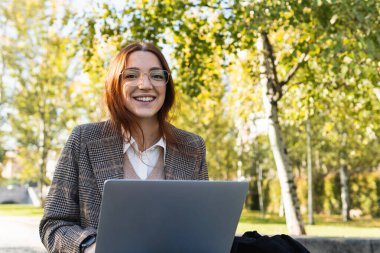 Bilgisayarda çalışırken kameraya gülümseyen gözlüklü güzel bir iş kadını. Parkta oturan genç, profesyonel kızıl saçlı kadın.