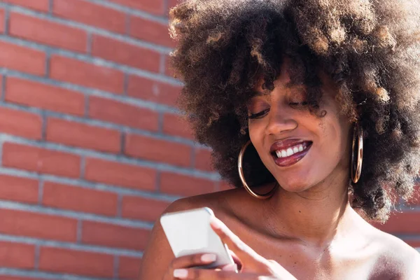 Sosyal medyayı kontrol eden sevimli Afro-Amerikan kadın dışarıda akıllı telefon tutuyor. Beyaz gülüşü, uzun afro saçı ve tuğla duvarda büyüleyici makyajı olan Afrikalı etnik kadın yüzü.