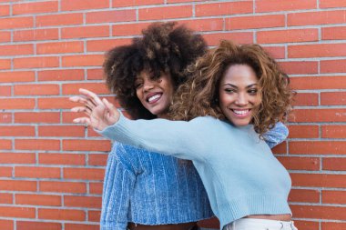 Afro saçlı, gösterişli makyajlı ve mavi kazaklı gülümseyen Afro-Amerikalı arkadaşların yan görüntüsü. Afrikalı doğal kızların güzel portresi tuğla duvarda eğleniyor.