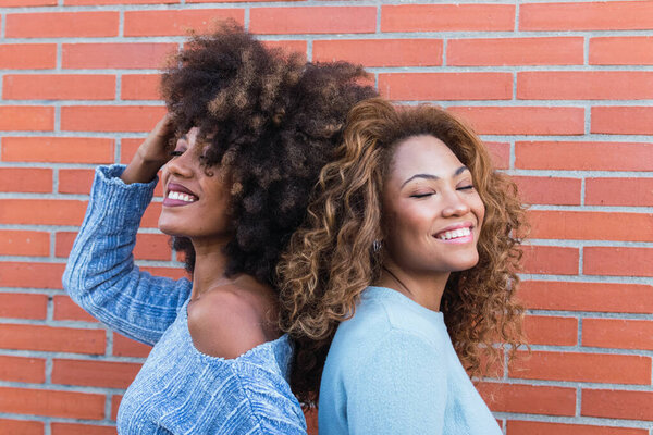 Side view of smiling african american women with long afro hair, glamour makeup and blue jumper posing in brick wall. Portrait of african natural girls standing back to back with eyes closed