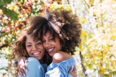 İki Afro-Amerikan mutlu en iyi arkadaşının birbirine sarılıp parkta gülüşmesi. Kıvırcık saçlı, sevgi dolu gülümseyen ve sıcacık sarılan neşeli afro siyahi arkadaşlar.