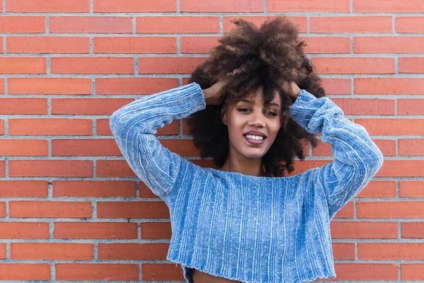 Çekici Afro-Amerikalı kadın uzun afro saçlarıyla gülümsüyor, göz kamaştırıcı makyaj yapıyor ve duvara dokunurken mavi kazak pozu veriyor. Güzel, siyah tenli, Afrikalı, etnik kadın yüzü.