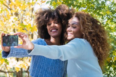 Selfie çekerken gülümseyen iki Afro-Amerikalı kadının portresi. Kıvırcık saçlı, mavi kazaklı ve selfie çekerken gülümseyen Afro siyahi arkadaşlar. Bulanık arkaplan ve dişçi kavramı