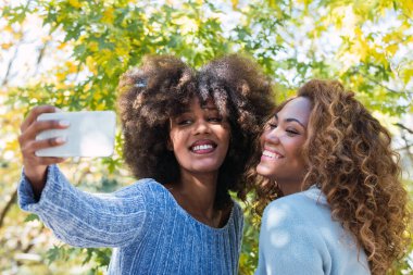 Selfie çekerken gülümseyen iki Afro-Amerikan bayan arkadaş. Kıvırcık saçlı, mavi kazaklı ve selfie çekerken gülümseyen Afro siyahi arkadaşlar. İlişki, teknoloji ve dişçi kavramı