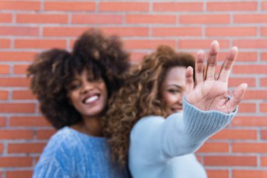 Uzun afro saçlı ve mavi kazaklı gülümseyen Afro-Amerikan kadınları kameraya doğru çekiliyorlar. Tuğla duvarda eğlenen Afrikalı doğal kızların güzel portresi. Eline odaklan.