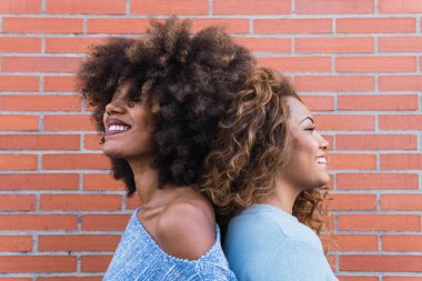 Uzun afro saçlı, parlak makyajlı ve mavi gömlekli, duvara poz veren gülümseyen Afro-Amerikan kadınları. Afrikalı doğal kızların güzel portresi kapalı gözlerle sırt sırta dururlar.