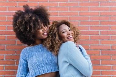 Gülümseyen Afro-Amerikan kadınları uzun afro saçlı, göz kamaştırıcı makyajlı ve mavi gömlekli tuğla duvarda poz veriyorlar. Afrikalı doğal kızların güzel portresi. Sarılırken eğleniyorlar.
