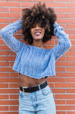 Çekici Afro-Amerikan mankeni, uzun afro saçlı, göz alıcı makyajlı ve mavi gömlekli. Tuğla duvardaki saçlarına dokunurken poz veriyor. Güzel, siyah tenli, Afrikalı, etnik kadın yüzü.