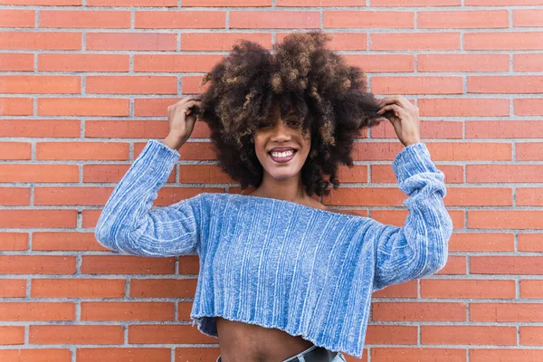 Uzun afro saçlarıyla gülümseyen Afro-Amerikalı bir kadın. Parlak makyajlı ve mavi gömlekli. Tuğla duvarda saçlarına dokunurken poz veriyor. Güzel, siyah tenli, Afrikalı, etnik kadın yüzü.