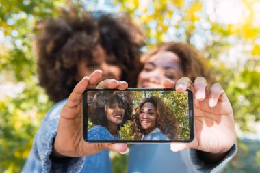 Neşeli, kıvırcık saçlı siyahi arkadaşlar ön kamerayla akıllı telefonla selfie çekiyorlar. Selfie çekerken gülümseyen iki Afro-Amerikalı kadın.
